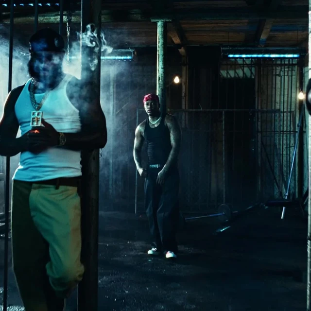Two stylish men in a dimly lit setting, one smoking, both wearing casual attire and jewelry, creating a moody and urban atmosphere.