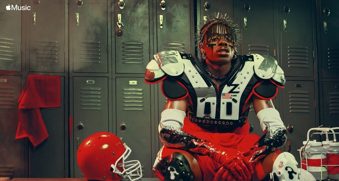 Man in football gear seated in locker room, red helmet on floor, water bottles; Apple Music logo in corner; studio lighting.