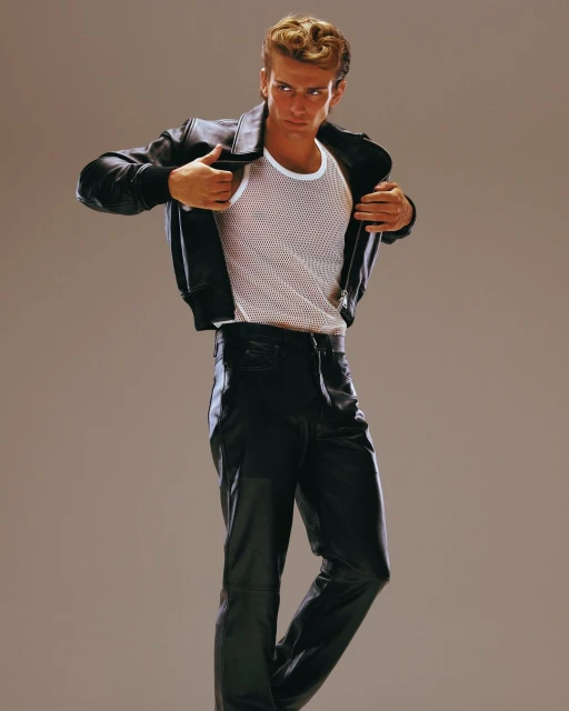 Man in black leather jacket and pants poses in a photo studio, wearing a white fishnet shirt. Fashion shot with neutral background.