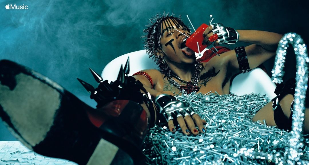 Man in tub filled with nails drinks from red container. He has face paint, spiked accessories, and intense expression against smoky blue backdrop.