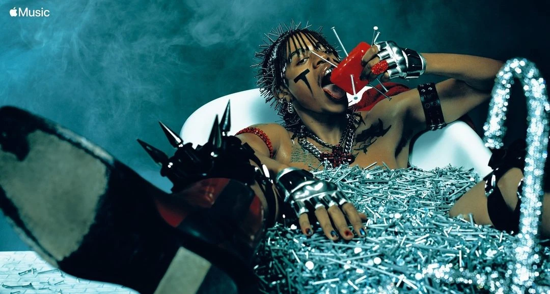 Man in tub filled with nails drinks from red container. He has face paint, spiked accessories, and intense expression against smoky blue backdrop.