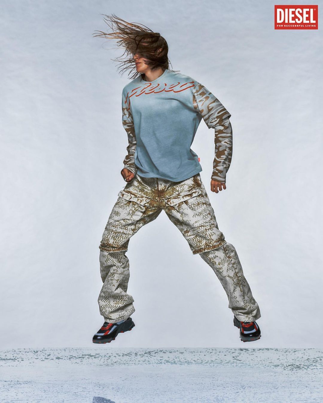 Person in mid-air wearing a Diesel t-shirt and patterned pants against a light backdrop, showcasing fashion and athletic movement.