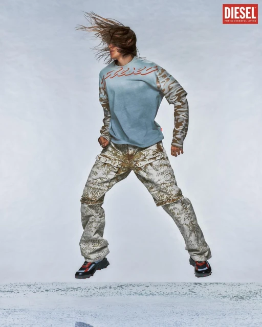 Person in mid-air wearing a Diesel t-shirt and patterned pants against a light backdrop, showcasing fashion and athletic movement.