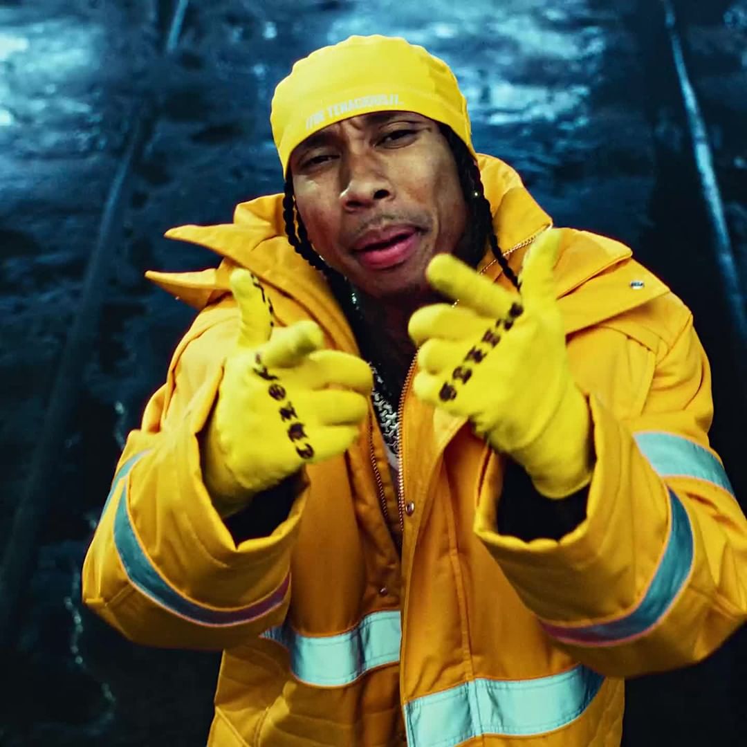Close-up of a male adult in a yellow outfit and hat, making hand gestures, set against an abstract blue and grey background.