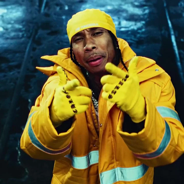 Close-up of a male adult in a yellow outfit and hat, making hand gestures, set against an abstract blue and grey background.
