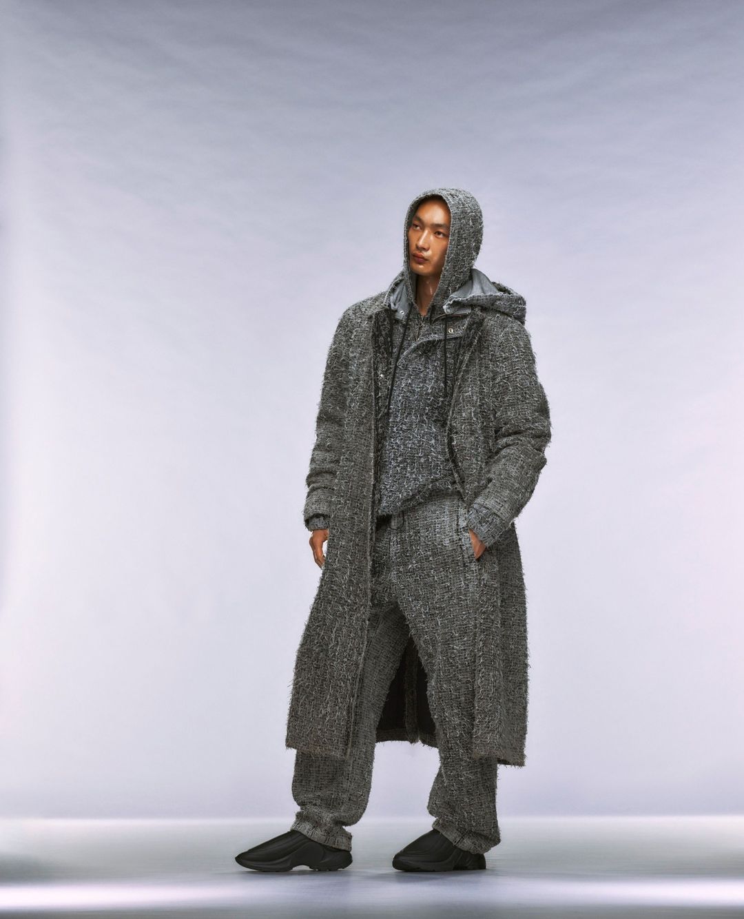 An Asian male stands in a photo studio wearing a grey hooded sweater, pants, and coat. The model has his hands in his pockets.