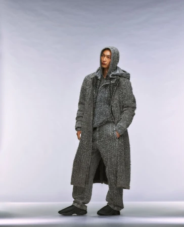 An Asian male stands in a photo studio wearing a grey hooded sweater, pants, and coat. The model has his hands in his pockets.