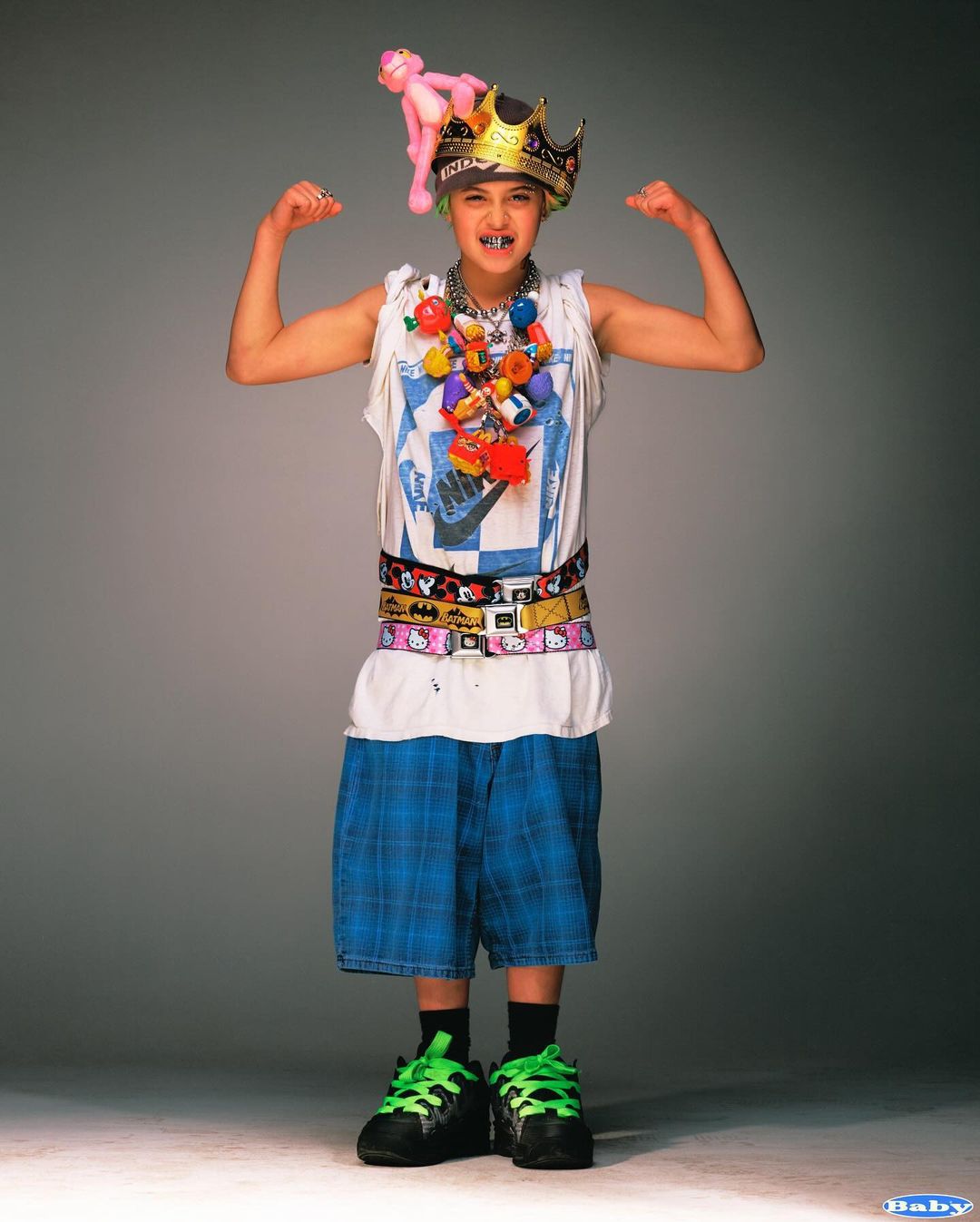 Young person strikes a fun pose indoors. They wear an oversized crown, a pink panther toy, and several belts. The individual stands in front of a grey background.