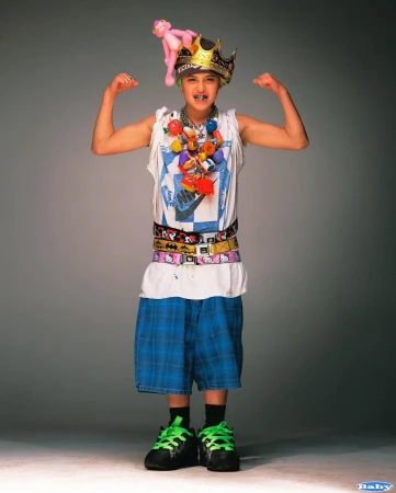 Young person strikes a fun pose indoors. They wear an oversized crown, a pink panther toy, and several belts. The individual stands in front of a grey background.