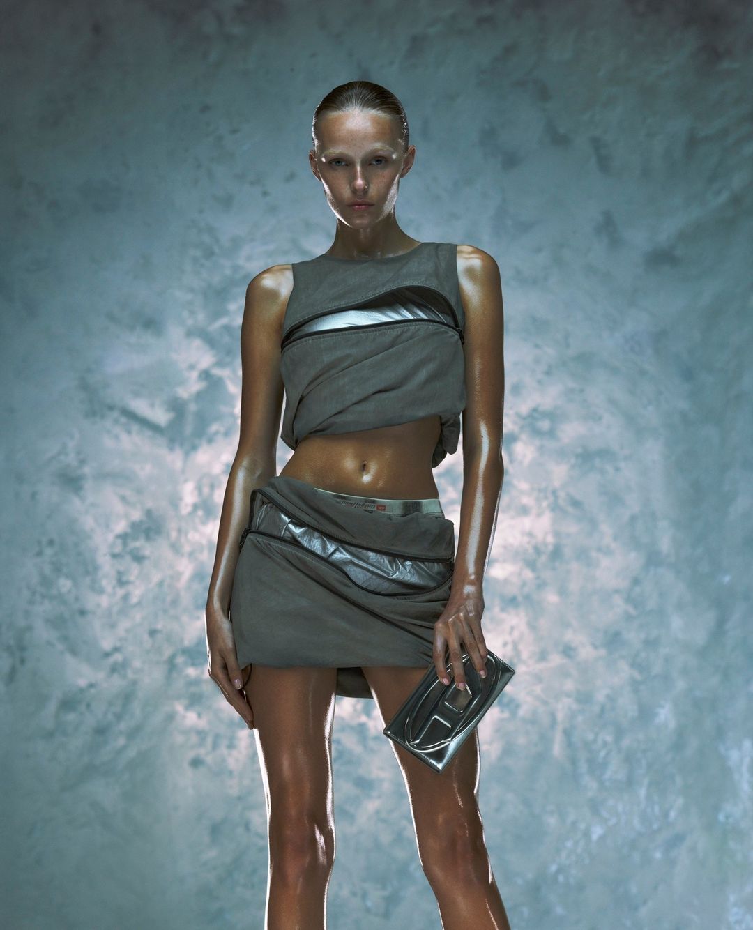 A lone adult female model, wearing a gray futuristic-style outfit and holding a clutch, is the primary focus against a textured background.