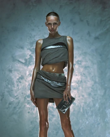 A lone adult female model, wearing a gray futuristic-style outfit and holding a clutch, is the primary focus against a textured background.