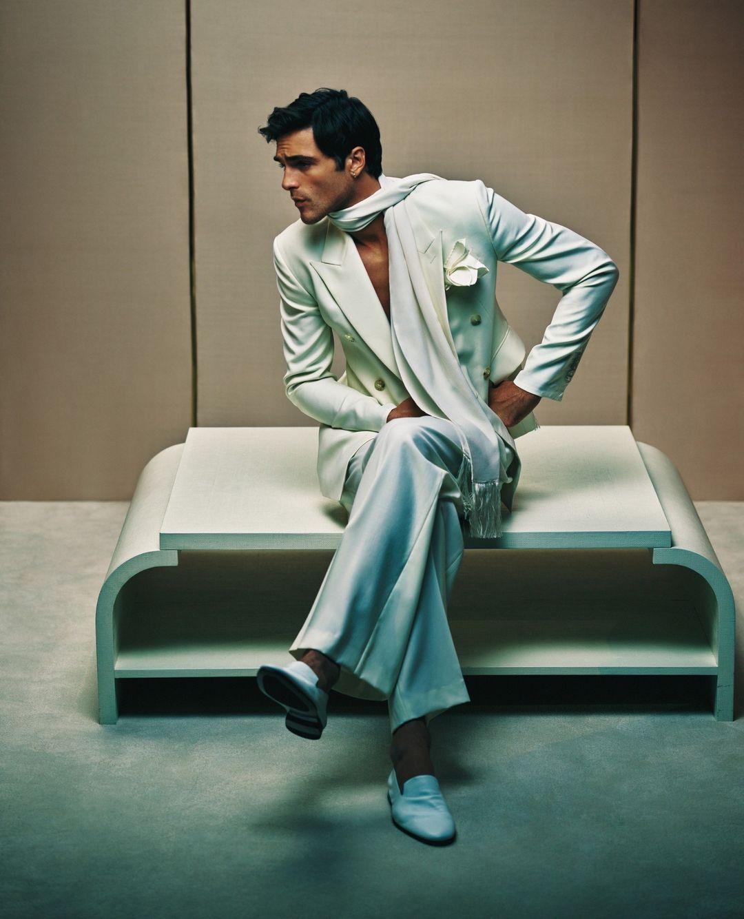 Man in a cream-colored suit and scarf seated on a platform, looking left. Fashion shot in front of a neutral studio backdrop.
