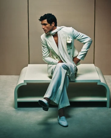 Man in a cream-colored suit and scarf seated on a platform, looking left. Fashion shot in front of a neutral studio backdrop.