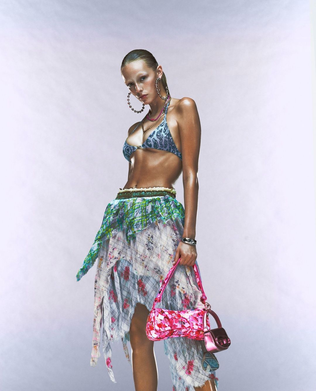A woman poses in a bikini top, wrap skirt, and jewelry, holding a pink purse against a white background in a fashion shot.