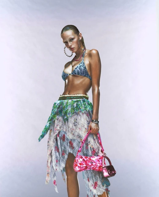 A woman poses in a bikini top, wrap skirt, and jewelry, holding a pink purse against a white background in a fashion shot.