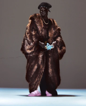 Full shot of a person in a brown fur coat, black pants, pink sneakers, and a black beret, holding an object in their hands.