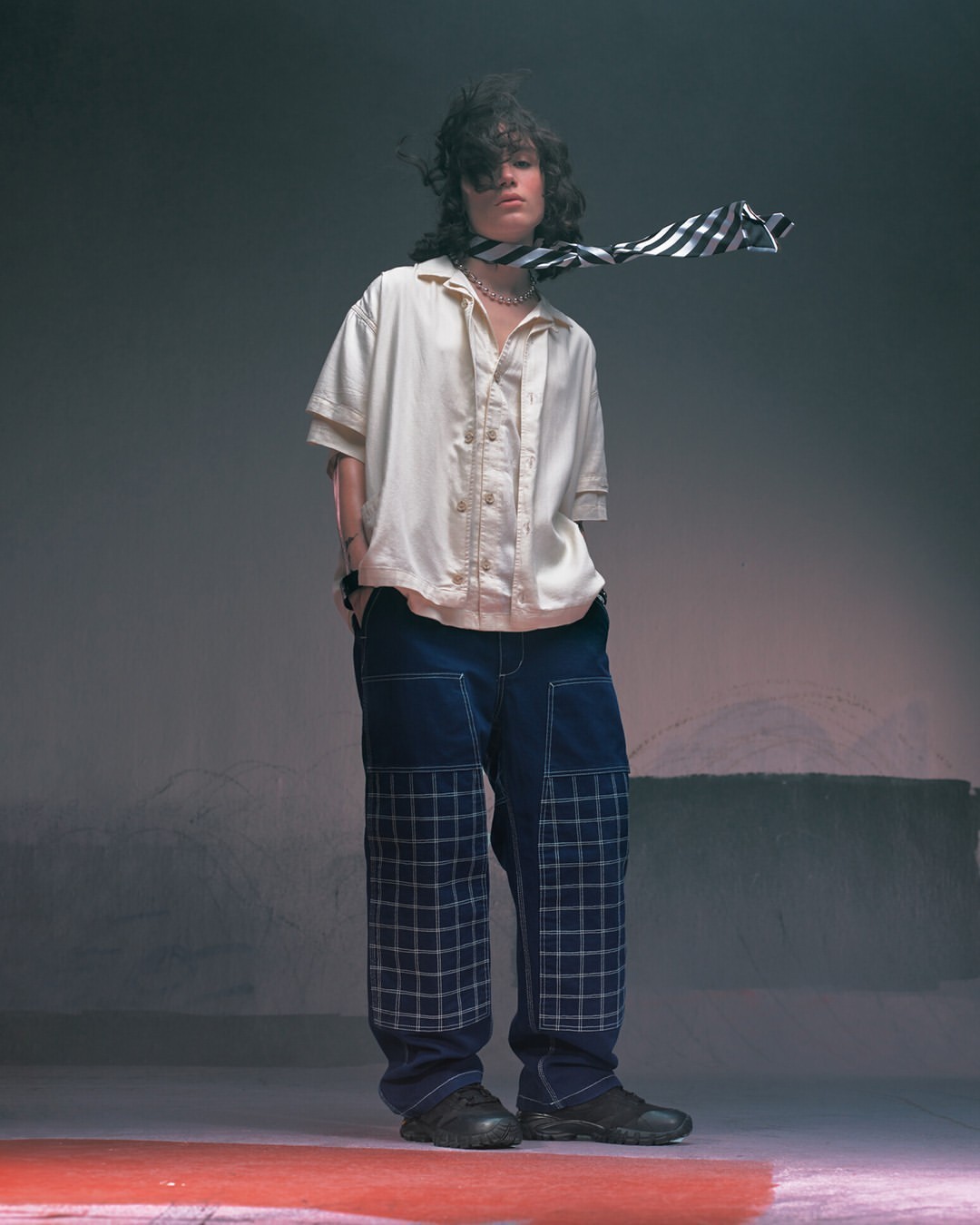 Person in cream shirt and blue jeans poses in a studio. Striped necktie adds flair to the fashion-forward look against a grey background.