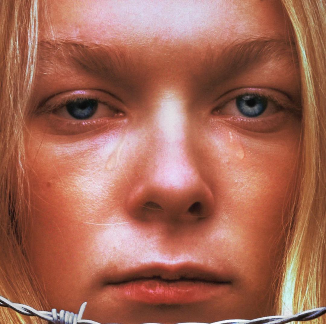 Close-up portrait of a tearful young woman with blonde hair and blue eyes near barbed wire with a shallow depth of field.