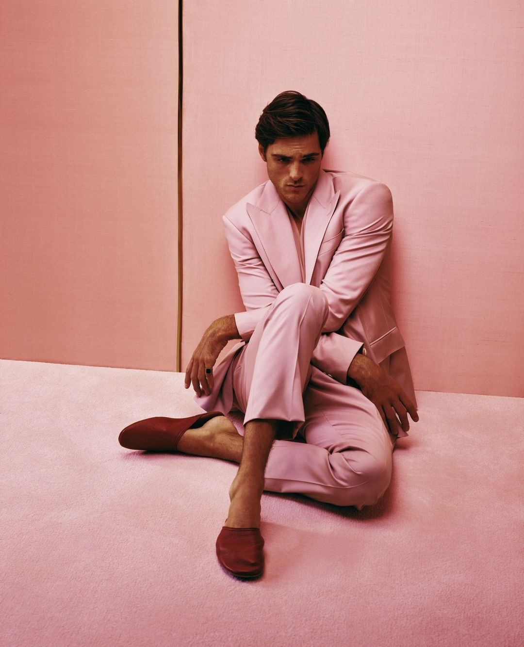 A relaxed man in a pink suit poses indoors against a pink backdrop, seated barefoot on a matching pink floor with red loafers.