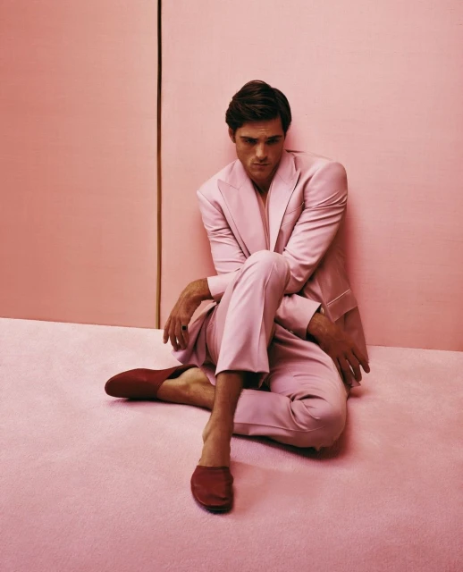 A relaxed man in a pink suit poses indoors against a pink backdrop, seated barefoot on a matching pink floor with red loafers.