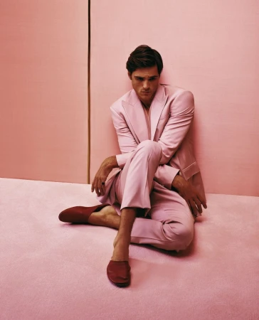 A relaxed man in a pink suit poses indoors against a pink backdrop, seated barefoot on a matching pink floor with red loafers.