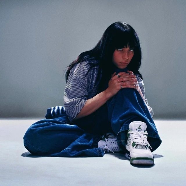 Young woman with dark hair sitting on the floor wearing oversized jeans and a striped shirt, posing in front of a grey background.