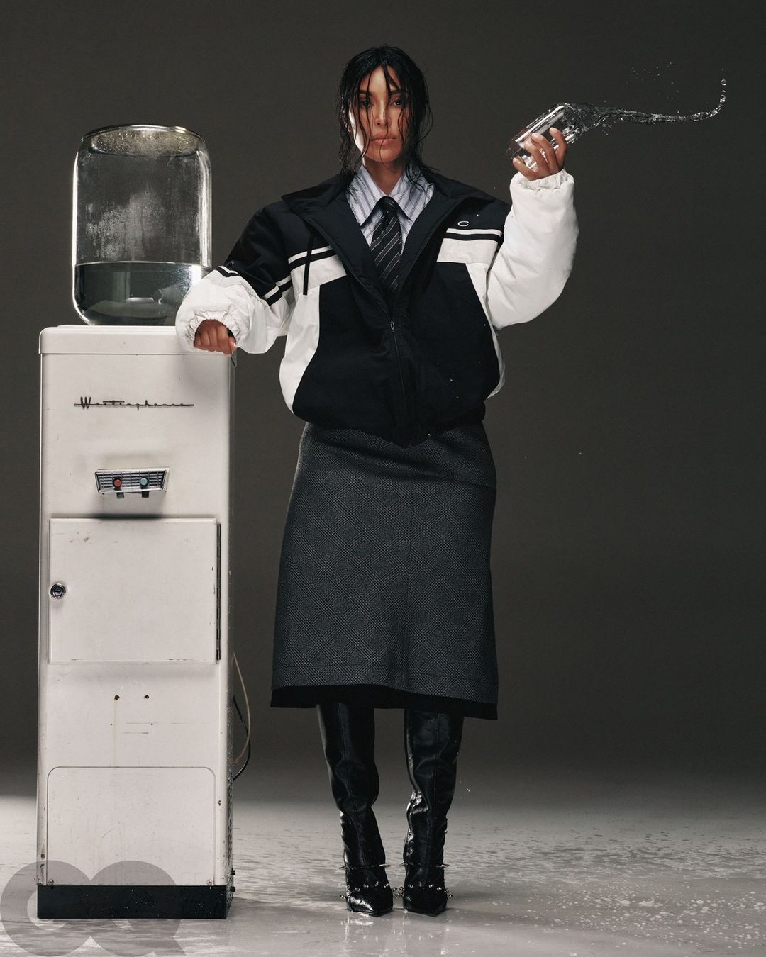 Adult female in an oversized jacket poses next to a water cooler, holding a glass with water splashing out in a fashion editorial.