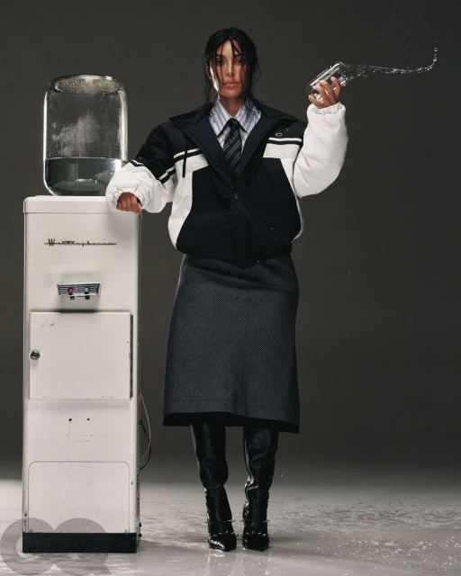 Adult female in an oversized jacket poses next to a water cooler, holding a glass with water splashing out in a fashion editorial.