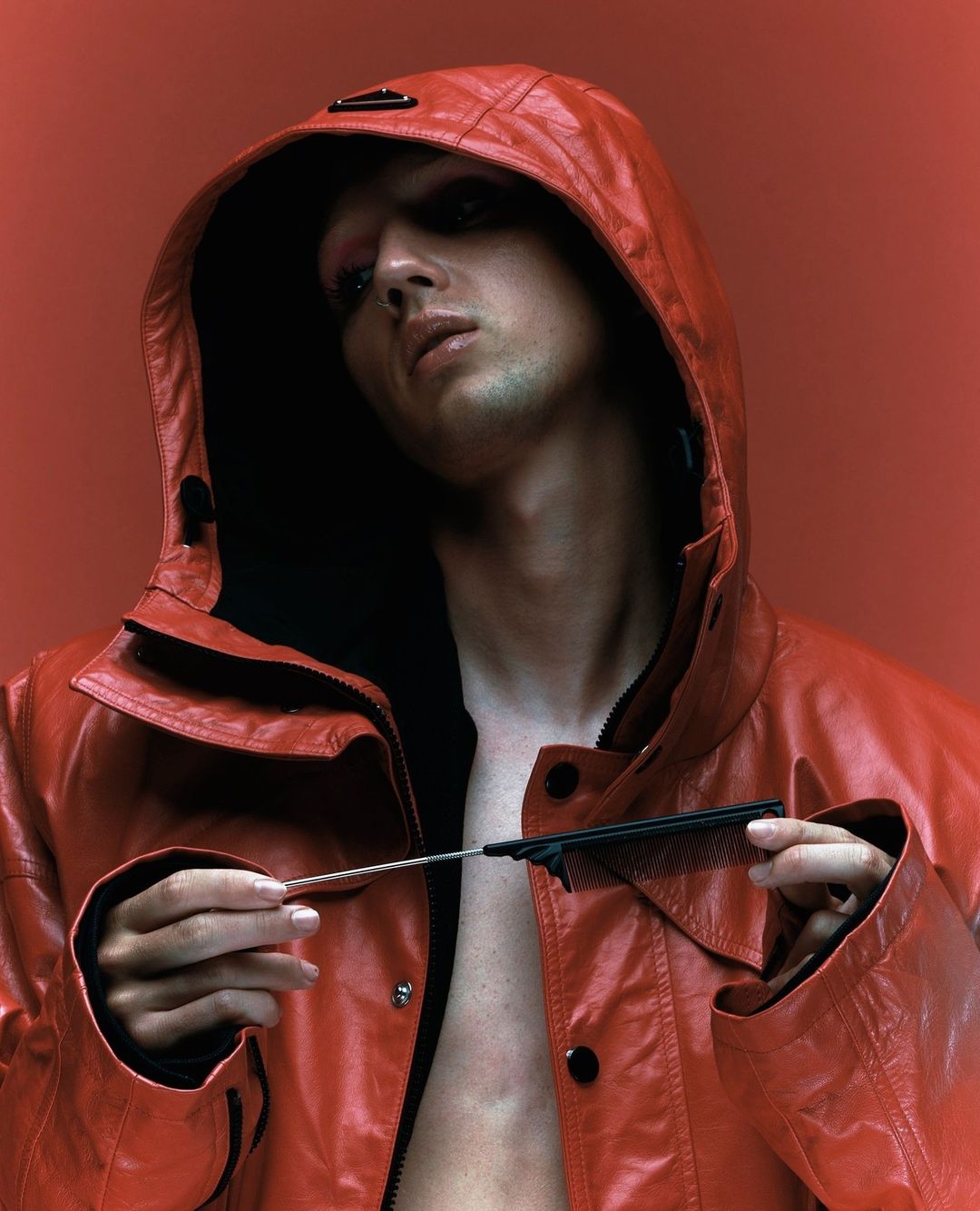 Close-up portrait of a young adult with a hooded red leather jacket, holding a comb, against a monochromatic red background in a photo studio.