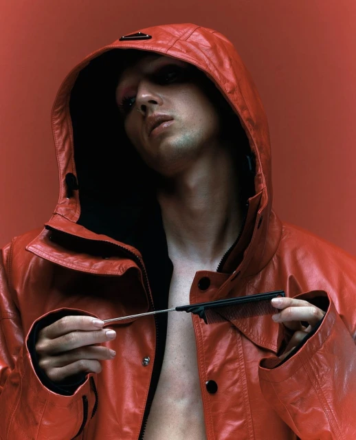 Close-up portrait of a young adult with a hooded red leather jacket, holding a comb, against a monochromatic red background in a photo studio.