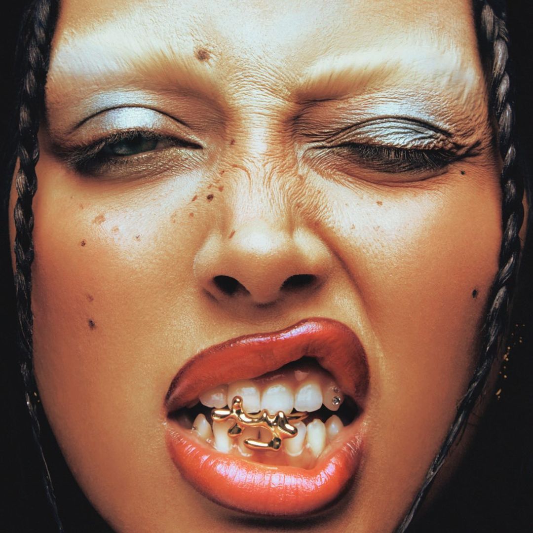 Close-up portrait of a young adult woman with dark braided hair, dramatic makeup, and a gold grill, against a dark background.