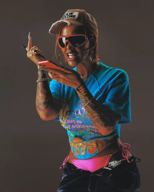 A tanned woman with tattoos, wearing sunglasses, a hat, and a T-shirt, is holding a phone while standing in front of a plain background.