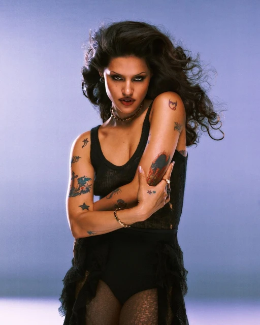 Portrait of a woman with dark wavy hair, wearing a black outfit and gold jewelry against a light blue backdrop in a studio shot.