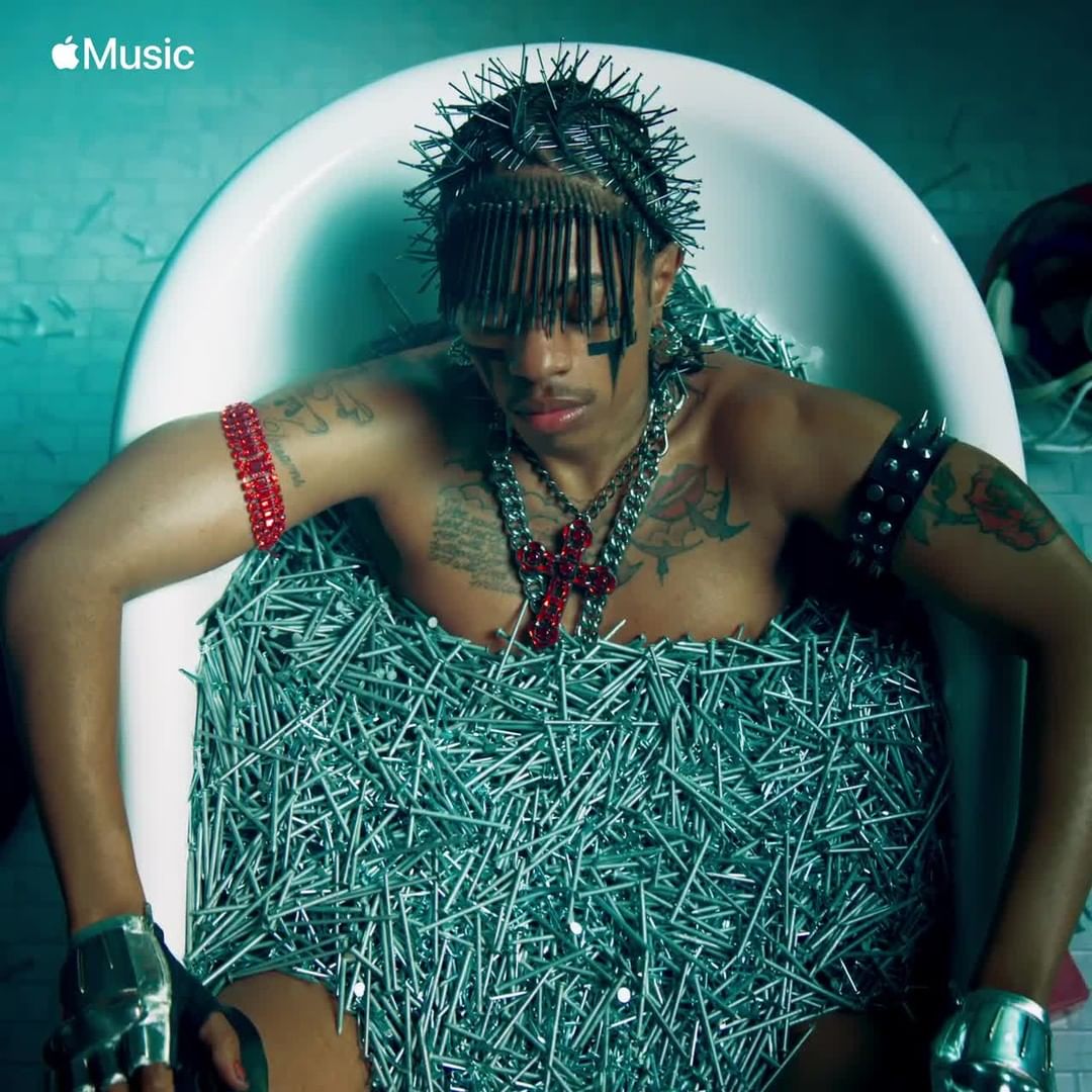 Adult male submerged in a bathtub filled with metal nails, adorned with edgy jewelry and decorative hair pins, in a music video.