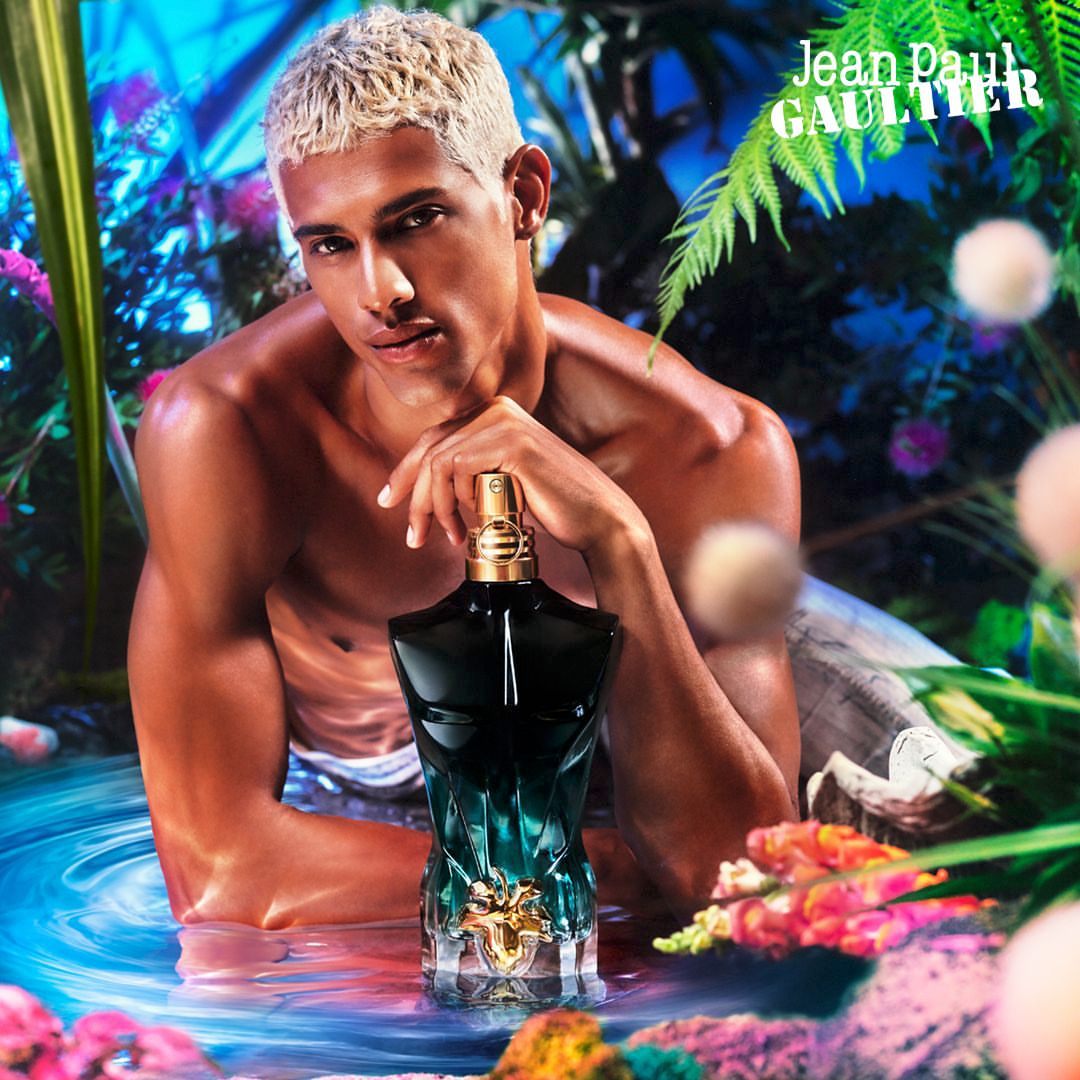 Masculine presenting person with platinum blonde hair, bare torso, and Jean Paul Gaultier perfume bottle in a floral aquatic environment.