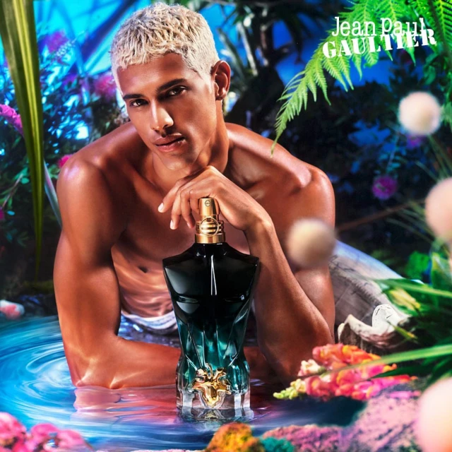 Masculine presenting person with platinum blonde hair, bare torso, and Jean Paul Gaultier perfume bottle in a floral aquatic environment.