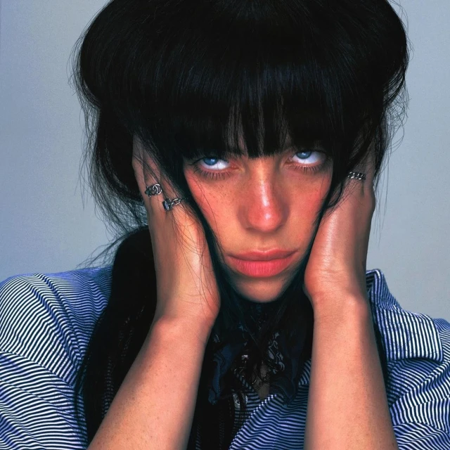 Headshot of a woman with black hair, blue eyes, wearing a striped shirt, holding her hands up to her face, and wearing rings.