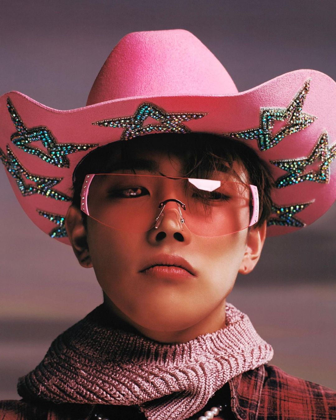 Close-up portrait of a person wearing a pink cowboy hat with star embellishments and pink glasses, posing in a photo studio setting.