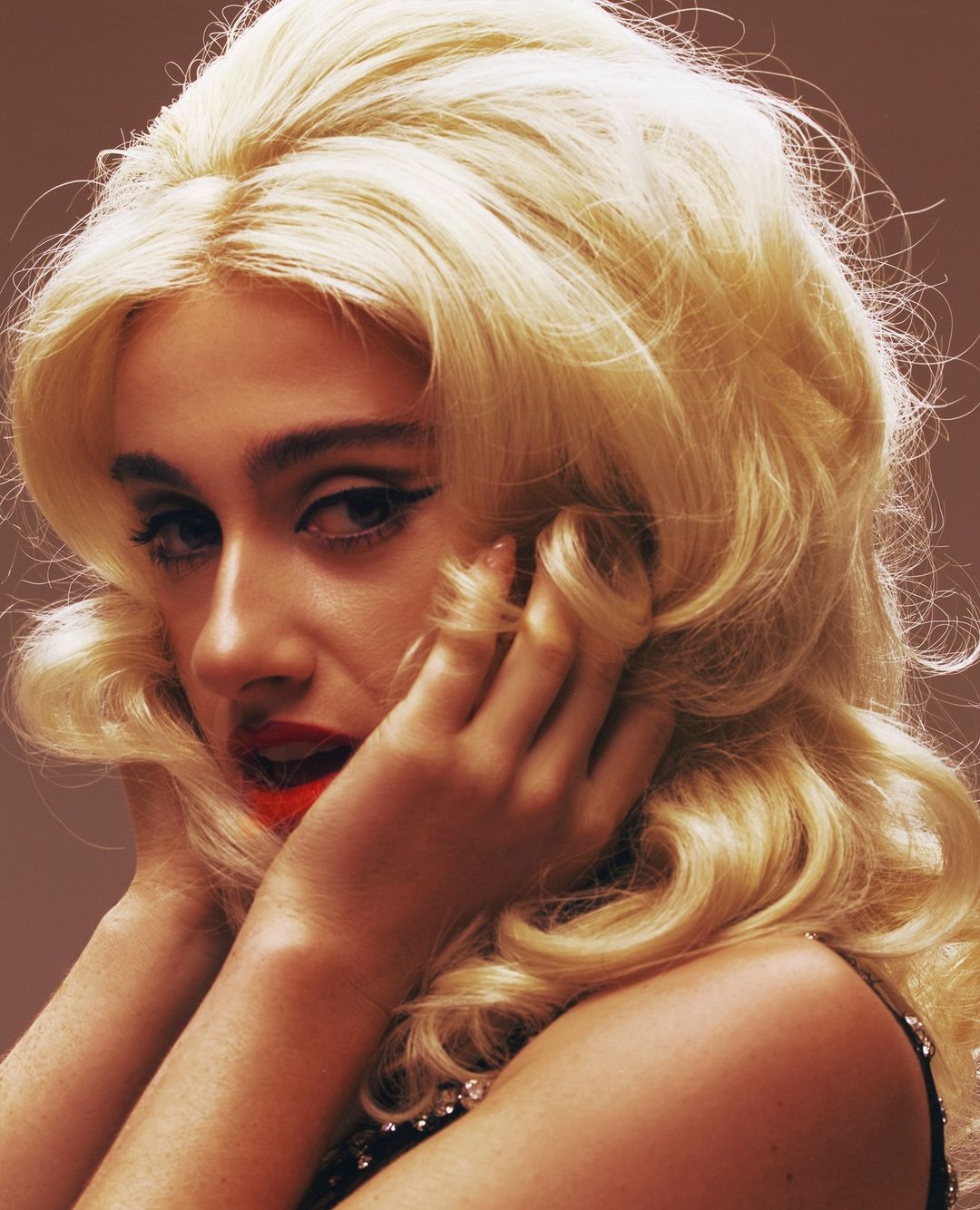 Close-up studio shot of a glamorous fair-skinned woman with big blonde wavy hair, dramatic eyeliner, and bright red lipstick.