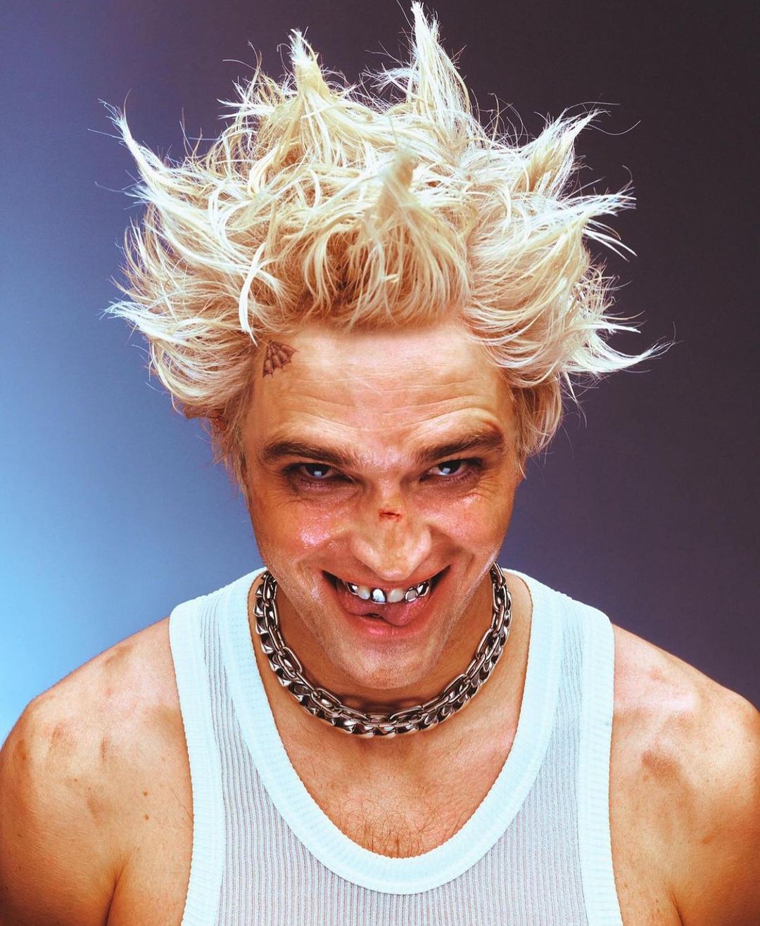 Close-up of a man with bleached spiky hair, a metal chain necklace, and a white tank top against a grey and blue gradient background.