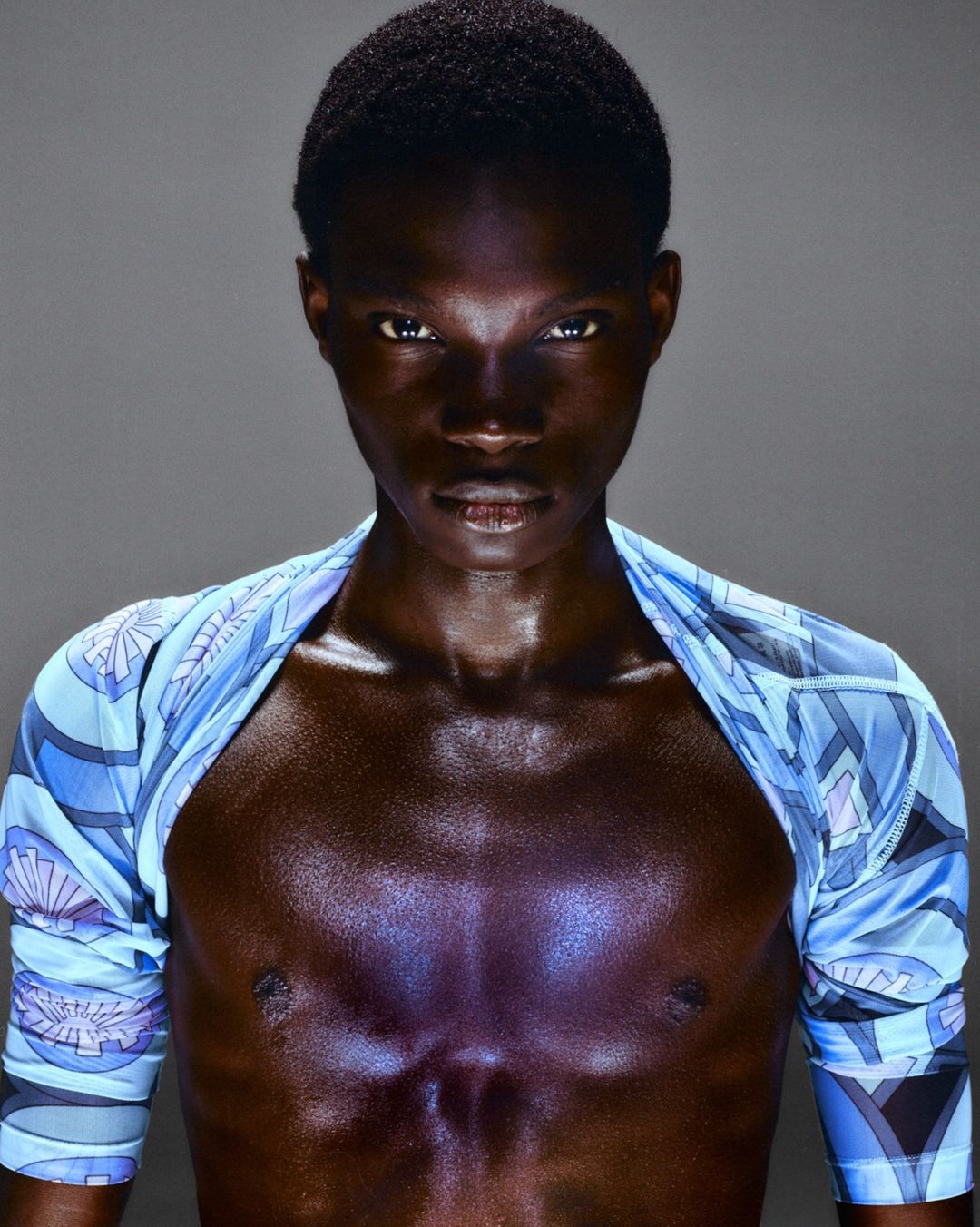 Portrait of a dark-skinned adult male looking at the camera with a patterned blue garment, medium shot, studio lighting on a gray background.