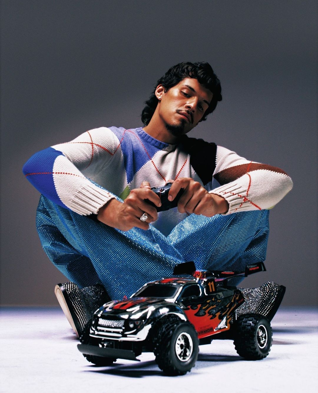 Man sitting, holding a remote control, with a toy car in front of him, wearing a patterned sweater and sparkling blue pants.