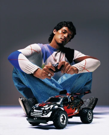 Man sitting, holding a remote control, with a toy car in front of him, wearing a patterned sweater and sparkling blue pants.