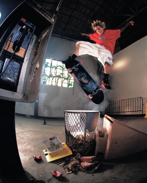 A young adult performs a skateboard trick in an urban indoor setting, jumping over a trash can near an old telephone booth.