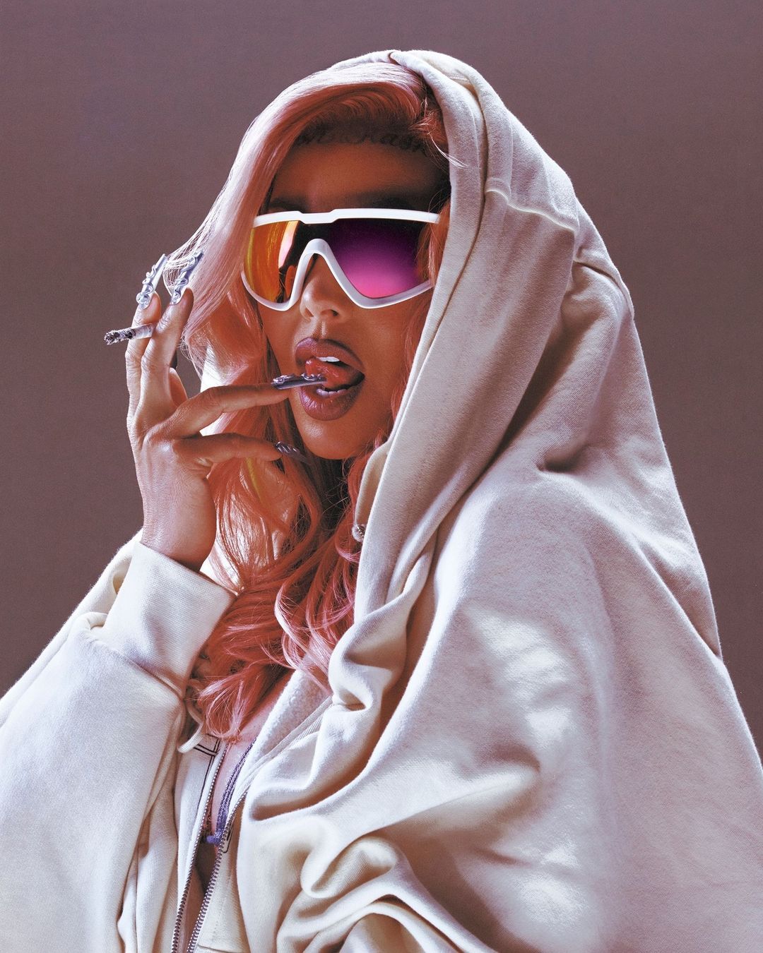 Woman with pink hair in a hoodie and futuristic sunglasses is biting a ring and holding a cigarette, posing indoors.