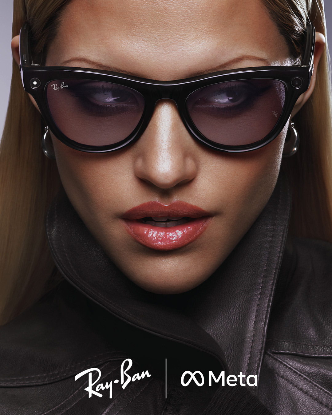 Close-up of a woman with sunglasses wearing a dark jacket, featuring Ray-Ban and Meta logos at the bottom, studio lighting.