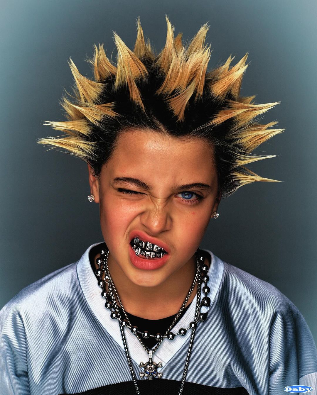 A young child with spiky hair, silver teeth grills, and multiple necklaces poses indoors, winking with one eye closed in a studio shot.
