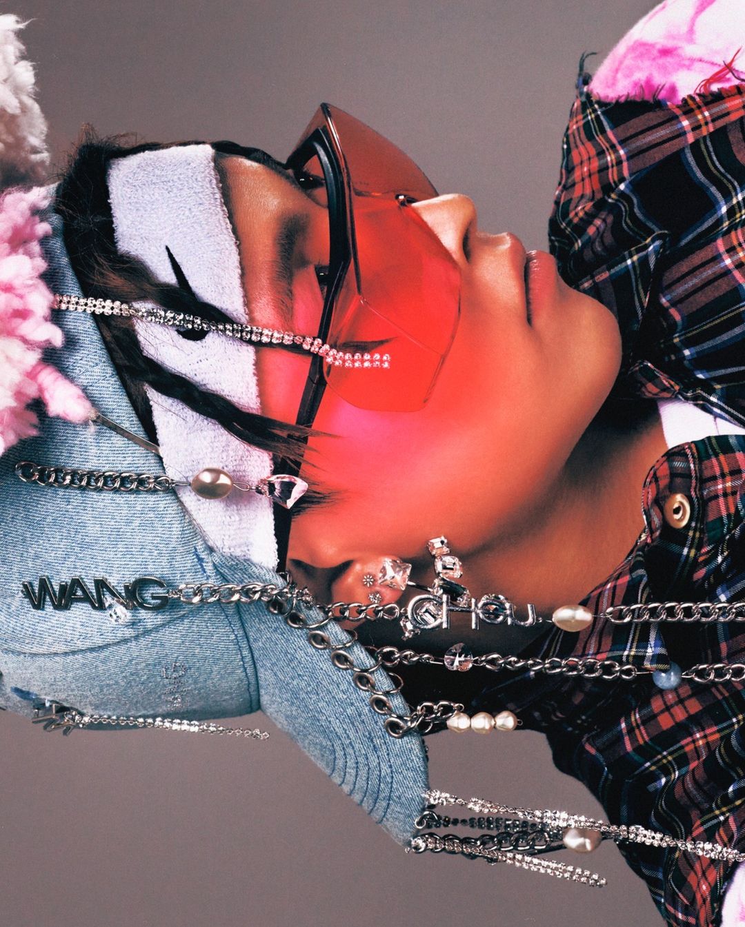 Portrait of a young adult wearing a plaid shirt, denim cap, and tinted glasses, accented with strands of pearls and rhinestone jewelry.