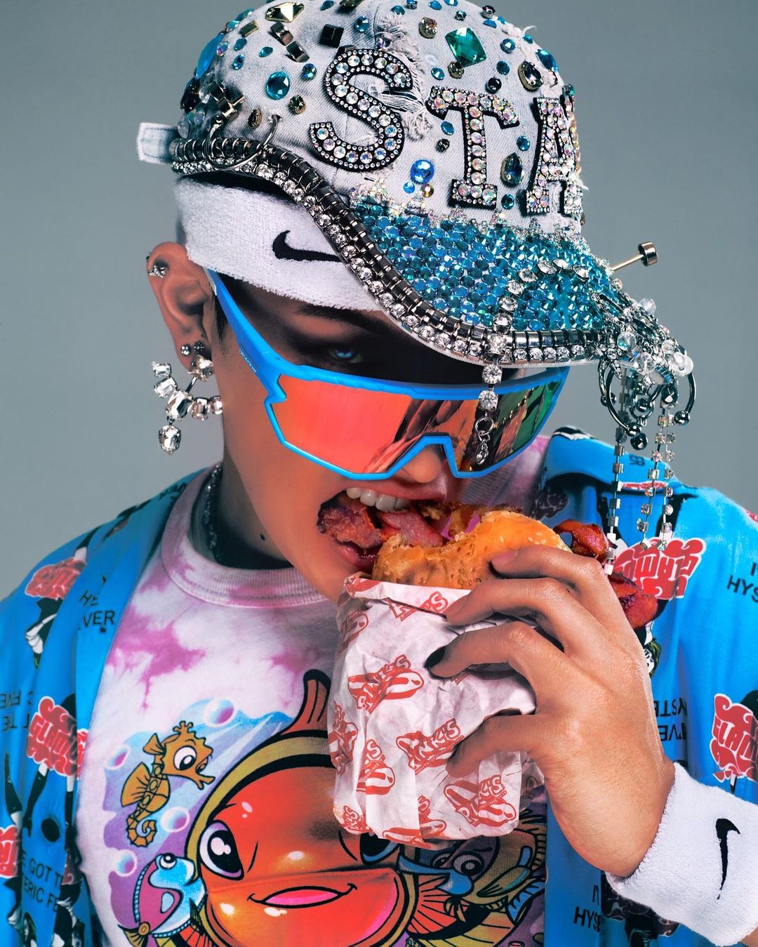 Adult wearing embellished baseball cap eating burger. Person in blue jacket, cartoon t-shirt, sunglasses, wristband, and jeweled earrings.
