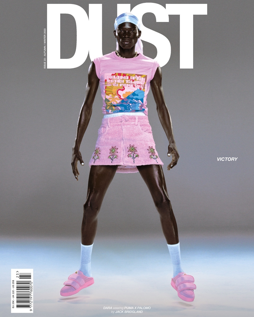 Dark-skinned male model posing in pink t-shirt, skirt, socks and sandals, wearing a durag and a necklace against a grey backdrop.